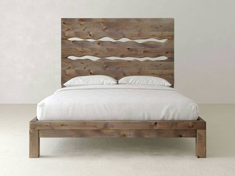 A James+James Live Edge Bed with a simple, rustic design sits in a minimalistic room. The handcrafted headboard features two wavy cut-out patterns, enhancing its unique artistic appeal. The bed is adorned with white bedding, including pillows and a neatly tucked duvet. The background is plain and light-colored.