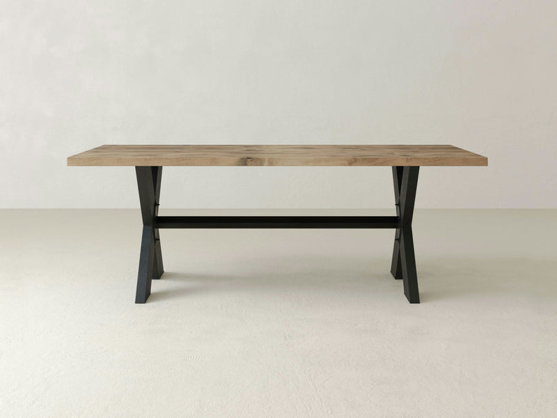 The X-Base Dining Table - Harvest Wheat by James+James features a minimalist wooden design with a light brown rectangular top and black metal legs. The table is showcased in an empty room with a plain off-white background and floor.