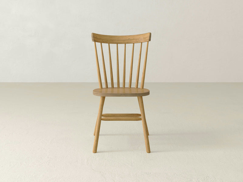 The Rustic Windsor Dining Chair - Harvest Wheat by James+James is a wooden chair with a round seat and a backrest featuring vertical support slats. It has four legs connected by horizontal support bars, all showcasing a natural, light finish. The chair is displayed against a plain white background.