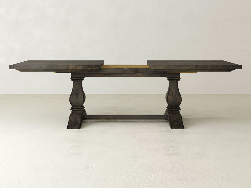 The Heirloom Expandable Dining Table by James+James, showcased with its leaves extended and finished in a dark hue, exemplifies rustic elegance. This wooden table features two rectangular support legs connected by a horizontal stretcher and boasts a visible central support section, all set against a plain light-colored wall.