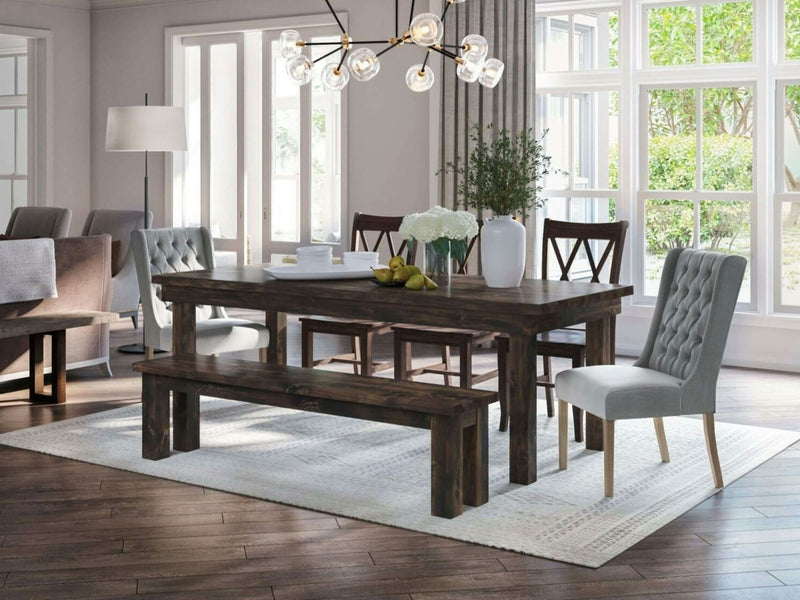 A modern dining room featuring a dark wooden table paired with the James+James Farmhouse Bench in Tobacco and upholstered chairs. A chandelier with globe lights hangs above, casting a warm glow. Large windows allow natural light to flood the room. The decor includes a vase of flowers, books, and a bowl of lemons on the table, all set atop a white rug.