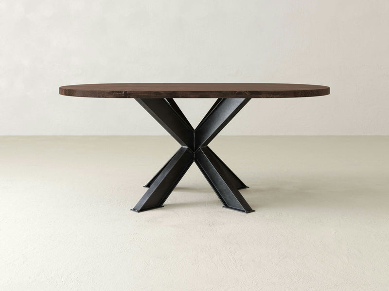 The Shiloh Oval Dining Table - Tobacco by James+James features a dark brown top and black metal X-shaped legs, set on a light-colored floor with a plain light beige wall in the background.