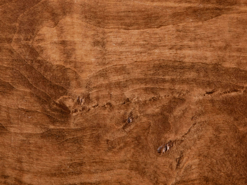 A close-up of the Tuscany on Maple Sample from James+James showcases a wooden surface with a natural brown grain pattern. The texture reveals subtle variations in color and grain direction, accentuating the organic lines and knots characteristic of wood.