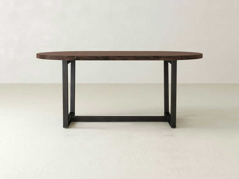 The Trapezoid Oval Dining Table - Tobacco by James+James features a modern oval-shaped wooden tabletop with a dark tobacco finish, supported by two black metal legs that form a rectangular frame at the base. The table is showcased against a plain, light-colored background.