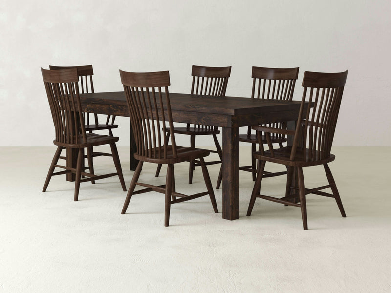 The James & James Farmhouse & Eleanor Dining Set includes a rectangular dark wood farmhouse dining table, handcrafted in the USA, and is accompanied by six matching Tobacco Eleanor Dining Chairs. The chairs feature high, slatted backs and are neatly arranged around the table in a minimalist, well-lit room with a light-colored floor and plain white walls.