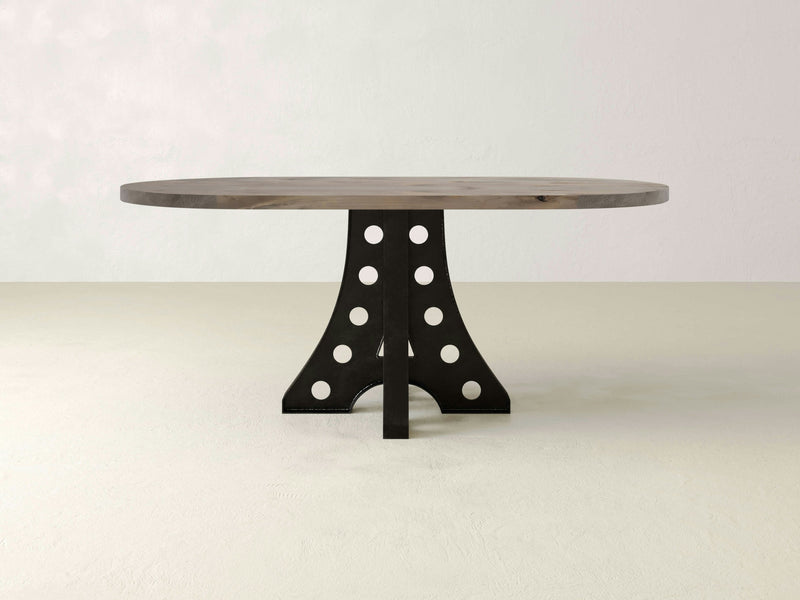 An Amelia Oval Dining Table in Barn Wood from James+James, characterized by a sturdy dark base with cutout circles arranged symmetrically, is placed in a minimally decorated room with light-colored walls and floor.