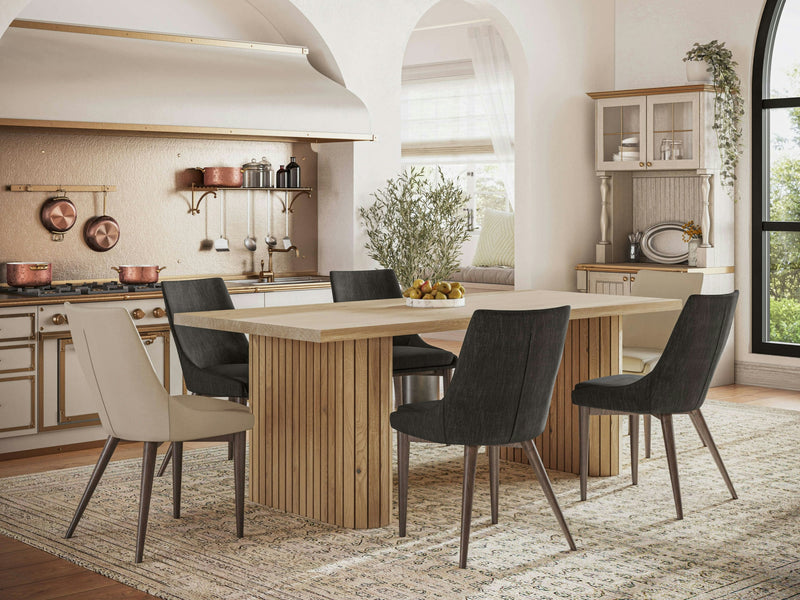 A modern kitchen with a Fern Dining Table - Satin by James+James, surrounded by six mixed gray and beige chairs. The kitchen features white cabinetry, copper cookware, and an arched window streaming light. A plant centerpiece and a fruit bowl adorn the table, adding a homey touch.