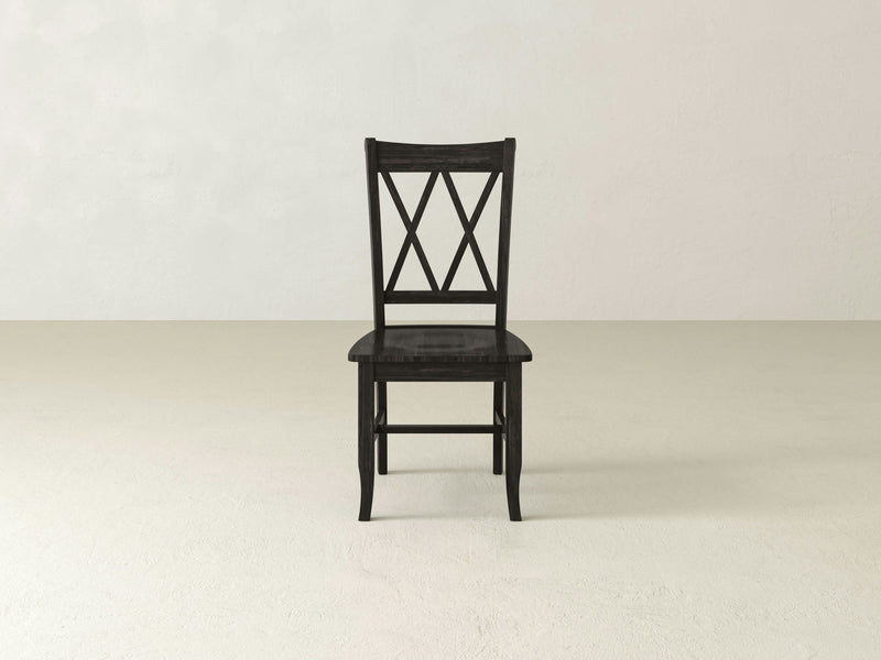 The Double X-Back Dining Chair by James+James, a black wooden masterpiece featuring a tall backrest that highlights artisanal excellence, stands gracefully in the center of a light-colored, empty room. Its simple yet classic design with straight legs and a flat seat radiates timeless elegance.