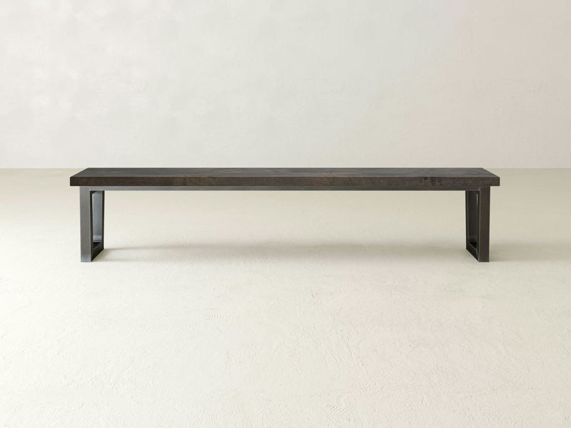 The Trapezoid Bench - Deep Grey by James+James features a minimalist design with clean lines and a rectangular seat, positioned against a plain, light-colored background. It has simple, sturdy trapezoidal legs on each end.