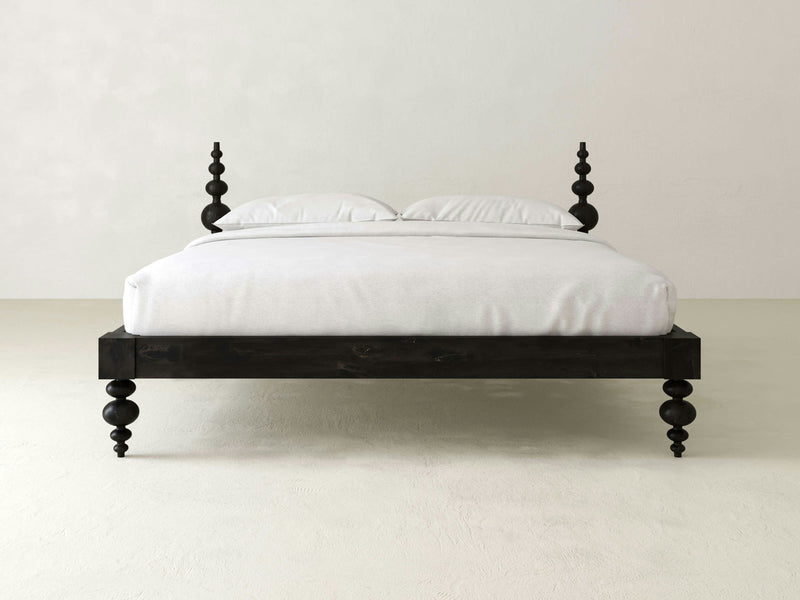 A minimalist bedroom features the Ophelia Bed - Charred Ember by James+James, showcasing an elegant dark wooden frame, intricately carved bedposts, and rounded legs. The bed is made up with white linens and two white pillows against a plain light-colored background.