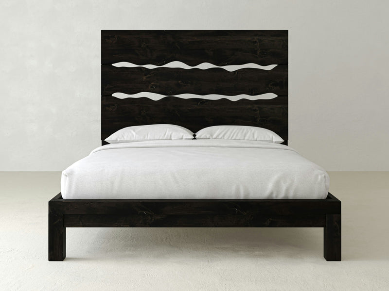 The Live Edge Bed - Charred Ember by James+James is a contemporary, dark wooden bed frame with a remarkable headboard showcasing wavy cut-out patterns. The bed is dressed in white sheets and pillows, accompanied by a neatly folded white blanket. It sits against a plain, light-colored wall, enhancing the minimalist ambiance.