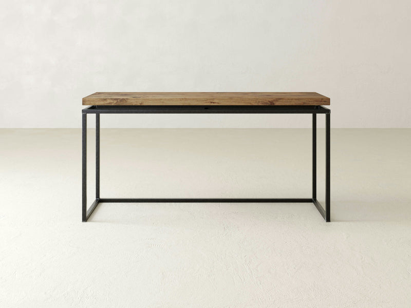 The Floating Top Sofa Table by James+James is a minimalist wooden piece featuring a thin rectangular tabletop paired with black metal legs. Its sleek and modern design highlights artisanal craftsmanship, boasting a simple yet elegant aesthetic. The table is set against a plain, light-colored wall and floor.