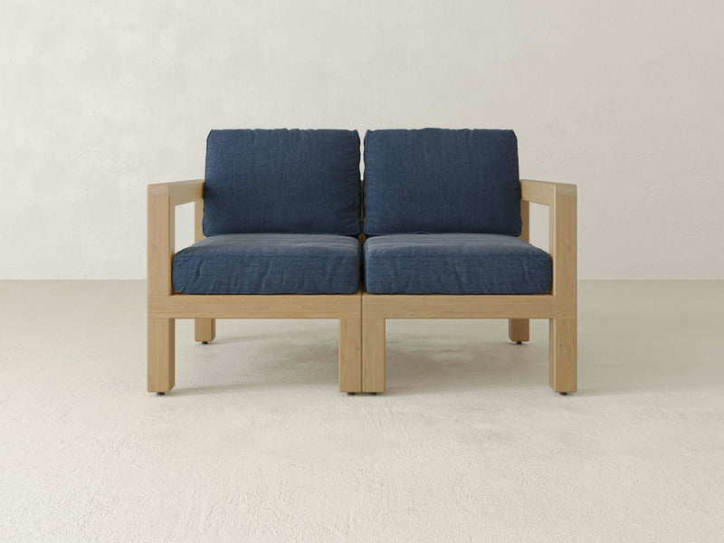 Presenting the Aria Outdoor Love Seat by James+James—a modern, minimalist wooden love seat with a light natural finish. This stylish piece features deep blue cushions on both the seat and back, offering a striking contrast against its light-colored wooden frame as well as against the plain white wall and floor background.