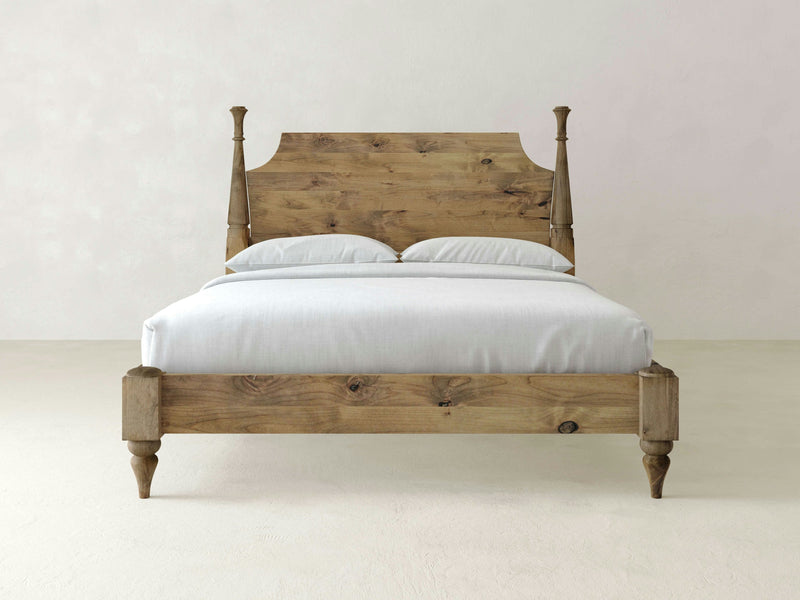 The Stella Bed - Harvest Wheat by James+James features a high, intricately carved headboard and four short, turned legs. Crafted from light-colored wood with prominent grain patterns, this bed is dressed in simple white bedding with neatly arranged pillows against the headboard set against a plain white wall background.