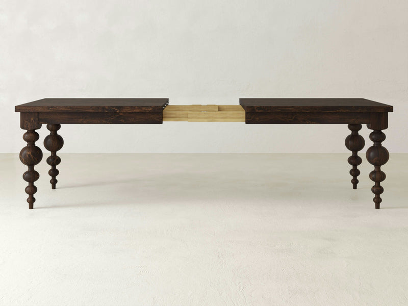 The Olivia Expandable Dining Table - Tobacco by James+James is shown in an extended position. The middle section, where the extension leaves would be added, is visible, revealing the lighter wood underneath. The table features dark ornate, turned legs and is placed in a minimalistic, neutral-colored room.