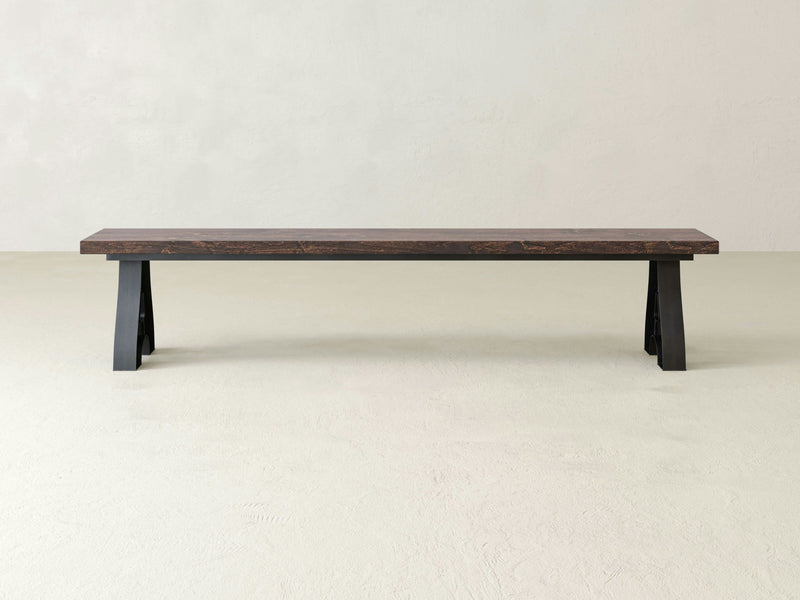 A long, minimalist Wishbone Bench - Tobacco by James+James, featuring a dark brown seat and black legs, is positioned against a plain, light-colored wall. The design is simple with its rectangular top and sturdy legs at each end, creating a modern and functional aesthetic.