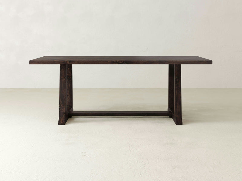 The James+James Cora Dining Table features modern aesthetics with its dark wooden rectangular top and sturdy trestle design. Showcasing two wide legs connected by a horizontal support beam, it stands elegantly against a plain, light-colored backdrop.