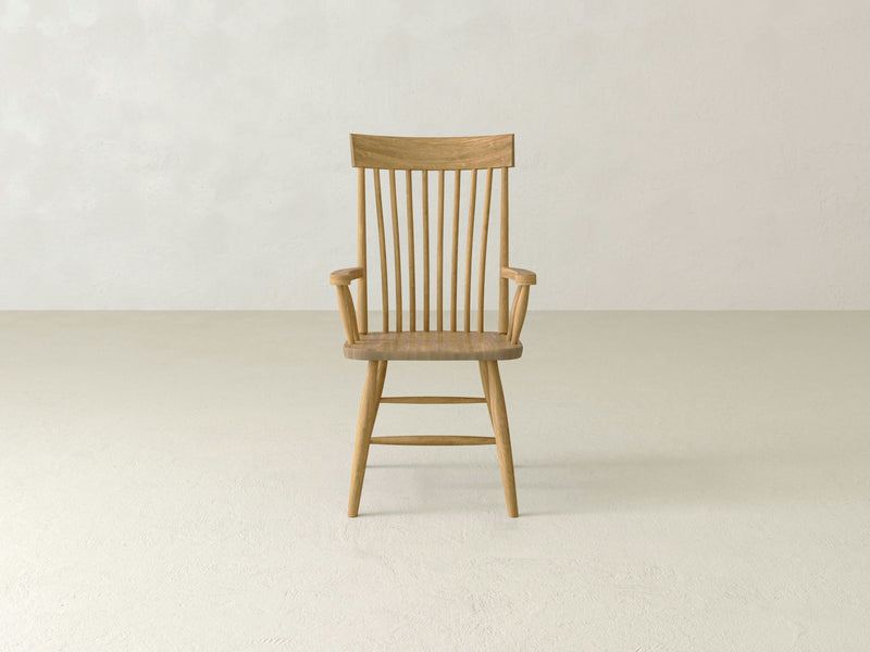 In the center of a plain, light-colored room stands an Eleanor Arm Dining Chair - Harvest Wheat by James+James. This wooden chair features a natural finish and boasts a high back with vertical slats and armrests. Positioned on four legs, it enhances the smooth, neutral-toned floor, contributing to the minimalist aesthetic of the space.